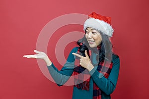 Pretty smiling asian woman in warm christmas sweater and santa hat pointing hand to present with red background for season