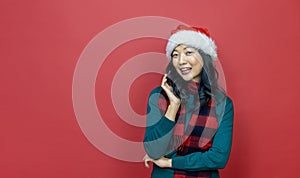 Pretty smiling asian woman in warm christmas sweater and santa hat on red background for season celebration concept