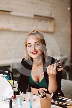 Pretty smiling beauty blogger dreamily looking in camera holding make up brushes in hand while recording new video for