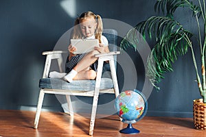 Pretty smiling blondy girl relaxing on a chair near the globe indoors at home with a tablet pc in her socks and jeans