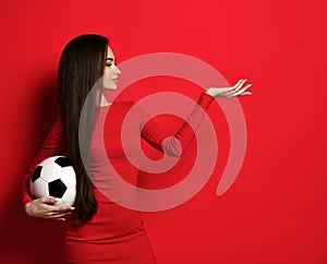 Pretty smiling brunette woman in tight red dress hold soccer ball in hand and pointing open palm hand with free text space on red