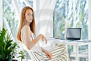 Pretty smiling businesswoman posing in a modern office sitting on her desk