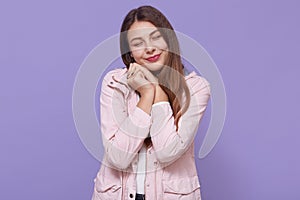 Pretty smiling charming young woman wearing pale pink jacket holding fists near cheeks isolated on lilac wall, keeping eyes closed