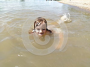 A pretty smiling girl of 6-7 years old is swimming in the water. Wet hair on the head. Laughing baby face. Serbia, Sava river