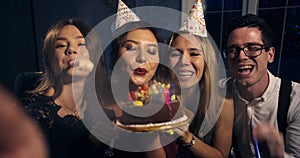 Pretty smiling girl blows out candles on a birthday cake surrounded by friends at a party. Around the atmosphere of the