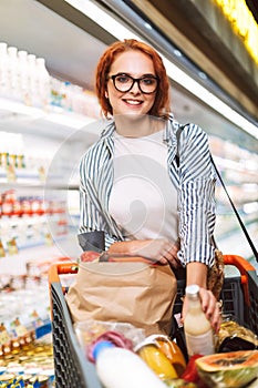 Pretty smiling girl in eyeglasses and striped shirt with shopping cart full of products happily looking in camera in