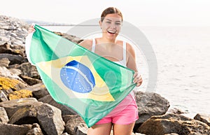 Pretty smiling girl holding Brazilian flag on the seashore on a sunny day