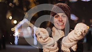 Pretty smiling girl in winter clothes, rejoicing, looking to camera, holding sparklers, spinning and dancing, standing in