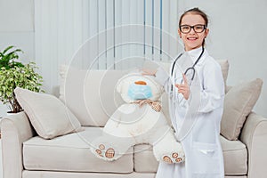 Pretty smiling little girl wearing doctor coat, stethoscope and glasses, standing, holding hand on the head of teddy