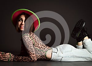 Pretty smiling playful young woman in wide-brimmed hat, leopard patterned longsleeve and jeans and hat is lying on floor