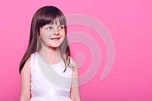 Pretty smiling small girl with dark hair, looking away with happy expression, wearing white dress, copy space for promotion,