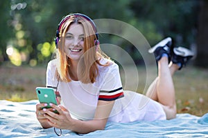 Pretty smiling teenage girl listening to music in pink earphones and using sellphone outdoors