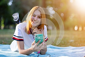 Pretty smiling teenage girl with red hair using sellphone outdoors in park