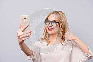 Pretty smiling woman with blonde hair in spectacles taking selfie and showing thumb-up