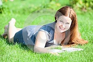 Pretty smiling woman lying on green grass with book