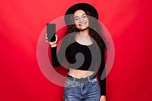 Pretty smiling woman smartphone screen and showing her finger at phone isolated on red background