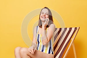 Pretty smiling woman wearing striped swimming suit sitting on deck chair isolated over yellow background talking on smart phone