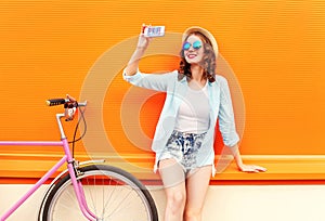 Pretty smiling young woman using taking self portrait on smartphone with retro bicycle over colorful orange