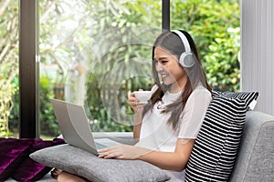 Pretty smiling young woman wear headphones sitting on sofa holding a cup of coffee and usinging on tablet