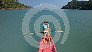 Pretty sportive blonde lady in stylish swimsuit sails on pink plastic canoe along azure ocean bay at exotic resort.