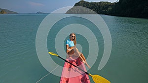 Pretty sportive blonde lady in stylish swimsuit sails on pink plastic canoe along azure ocean bay at exotic resort.