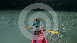 Pretty sportive blonde lady in stylish swimsuit sails on pink plastic canoe along azure ocean bay at exotic resort.