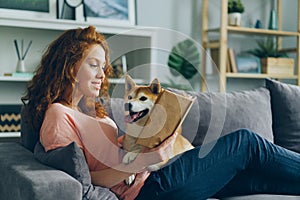 Pretty student reading book in apartment smiling and petting adorable dog