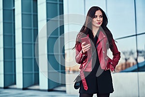 Pretty styilish woman in red jacket is standing next to glass building and chating with someone by mobile phone