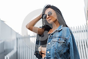 Pretty stylish young hipster woman in a trendy black t-shirt in sunglasses in a vintage denim jacket posing outdoors