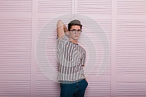 Pretty stylish young man hipster in fashionable glasses with a hairstyle in a trendy striped t-shirt stand near a vintage wooden