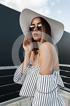 Pretty stylish young woman in fashionable summer dress in vogue sunglasses in vintage straw hat with sexy lips posing outdoors in