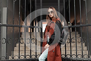 Pretty stylish young woman model in brown fashion trench coat in sunglasses with leather backpack posing near iron gate on street