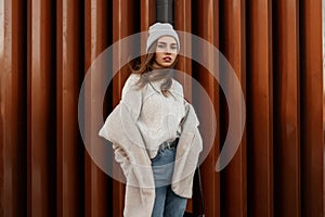 Pretty stylish young woman model in a fashionable knitted sweater in a knitted hat in an artificial fur coat in jeans stands near
