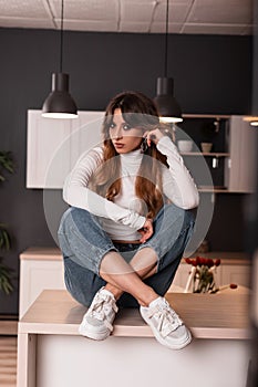 Pretty stylish young woman with red hair in trendy casual blue-white clothes posing sitting on a wooden table in the cook room.