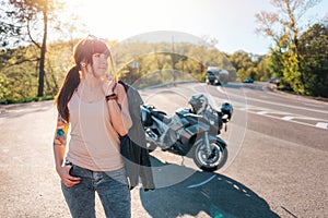 Pretty tattoed girl with a leather jacket poses against the background of a motorcycle by the road. Motorcycle local