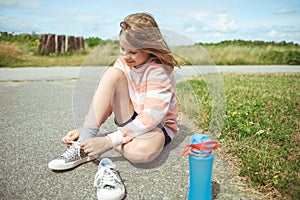 Pretty teen girl smiling and sitting on ground tie shoelaces at summer day