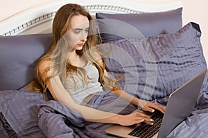 Pretty teen woman using her laptop in bed. Beautiful girl in glasses working on laptop in bedroom