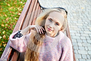 Pretty teenage girl 14-16 year old with curly long blonde hair in the green park on the bench in a summer day outdoors. Beautiful