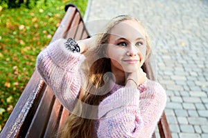 Pretty teenage girl 14-16 year old with curly long blonde hair in the green park on the bench in a summer day outdoors. Beautiful