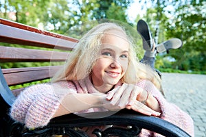 Pretty teenage girl 14-16 year old with curly long blonde hair in the green park on the bench in a summer day outdoors. Beautiful