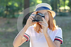 Pretty teenage girl with red hair taking picture with photo camera in summer park