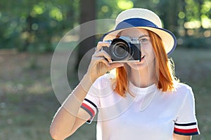 Pretty teenage girl with red hair taking picture with photo camera in summer park
