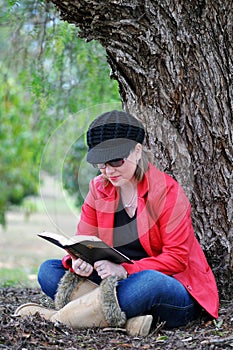 Pretty teenage girl studying holy bible beside huge tree in park