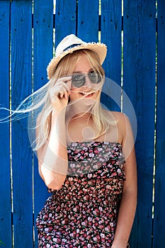 Cute teenage hipster girl smiling in straw hat, dress and sunglasses outdoor over blue wooden background.