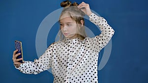 Pretty teenager girl looking to mobile camera for selfie photo on blue background. Young woman correcting hairstyle