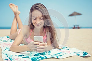 Pretty teenager girl using a smart phone lying on the beach with the sea and horizon in the background