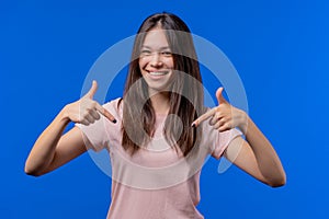 Pretty teenager pointing down to advertising area. Blue background. Young woman asking to click to subscribe below. Copy