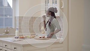 Pretty thoughtful african american woman with dreadlocks eating an apple in the kitchen looking in the window. Camera