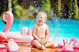 Baby sitting on the edge of the pool and smiling with pink flamingo lifebuoy