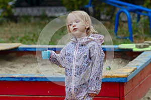 Pretty toddler girl wearing rain jacket playing with blue cup near the sandbox on the playground